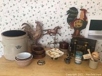 Photo showing entire lot including Pyrex bowls, wooden bowls, metal roosters, horse figurines, ceramic crocks and vintage tins on wooden surface against floral wallpaper.