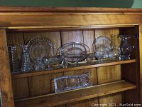 Full shelf view showing various clear glass pieces including serving dishes, pitchers, and decorative bowls arranged on two wooden shelves.