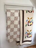 Two quilts hanging on rack, one floral patchwork and one with colorful leaf applique design on white background.