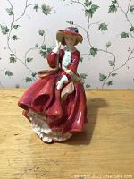 Front view of Royal Doulton figurine Top O' the Hill depicting woman in red dress and wide brown hat with blue ribbon