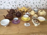 Overview of assorted tea pots, cups, and miniature tea sets arranged on wooden surface in front of a floral wallpapered background.