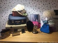 Photo of vintage travel suitcases, assorted hats including floral straw hat and furry cream hat on mannequin head, decorative boxes, framed pictures, and vintage clock under glass dome.