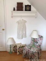 Lamps, floral upholstered chair, two floral needlepoint cushions, white hanging garment on hanger, small brown wooden jewelry box, and decorative clock on white shelf