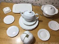 Photo showing variety of Wedgwood dinnerware and serveware pieces laid out on a wooden surface, includes plates, teapot, butter dish, covered serving dish, and decorative sphere.