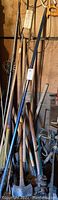 Various yard tools including axes, hatchets, metal poles, and a saw hanging and standing against wall.