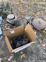 Overview of lot showing kettles, pot, bucket, sad irons in box, and metal lamp on grass with leaves