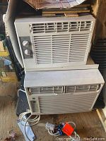 Two window air conditioning units stacked vertically in a shed, both white with visible control panels and power cords.