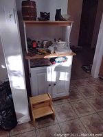 Kitchen cabinet with butcher block top, two shelves, and white double doors, shown with various items on shelves and countertop, accompanied by small wooden step stool on floor.
