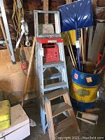 Metal step ladder with red box and blue snow shovel in background, along with a yellow and blue barrel holding tools