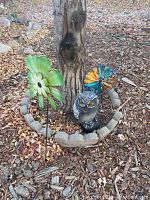Photo showing a large green metal flower yard stake and a realistic metal owl statue placed on mulch around a tree base, along with a colorful metal flower stake.