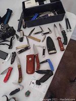 Photo showing assorted knives laid out on a white surface with knife cases and a black toolbox containing additional tools.