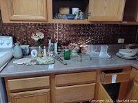 Wide view of kitchen counter showing various vintage kitchen items including vases, mold, salt and pepper shakers, trivets, placemats, and glassware.