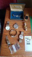 Seven wristwatches, one pocket watch, one loose watch head with band, and a bag of bezels displayed on wood surface alongside Washington watch box