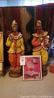Two handmade clay clown sculptures, approximately 24 inches tall, placed together with a framed clown artwork on the floor in front.