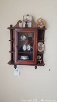 Full view of the dark wood wall mounted display case with glass door containing assorted Limoges porcelain items and two decorative framed pictures on top, with multiple shelves around the case displaying plates and porcelain pieces.