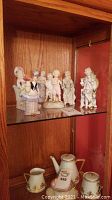 Display shelf with six porcelain figurines depicting people in historical dress and a porcelain tea set including teapot, creamer, and sugar bowl