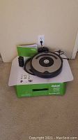 Overview of Roomba 677 on its original box along with charging dock, Virtual Wall barrier and manual on carpeted floor