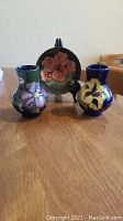Two Moorcroft vases, one with purple flower design and the other with yellow flower design, displayed with a floral plate on a stand in background.