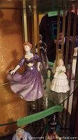Two Royal Doulton figurines displayed in glass cabinet; one woman in purple dress and a smaller girl in white dress holding basket