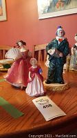 Three porcelain figurines on wooden table: clown at center, lady with fan at left, girl with book at front.