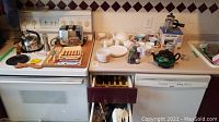 Wide shot showing toaster, kettle, two drawers with kitchen utensils and flatware, teapots and cups on counter.