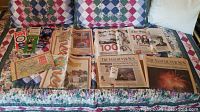 Vintage newspapers and printed materials laid out on a quilted surface, showing multiple newspapers and guide booklets related to Expo 86 and Millennium events.