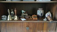 Shelf display of vintage and decorative collectibles including brass bells, a wooden cat figurine, ceramic bell, framed picture and wooden whistle