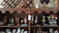Wide view of wooden shelving unit holding various vintage decorative items including ceramic plates, figurines, ceramic diner flower pot with flowers, wooden cat figurines, and black vase.
