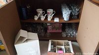 Full view of cabinet shelving with assorted glassware including hand painted mugs, clear small glasses, and boxed glass items.