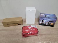 Four boxed or packaged items displayed on a wooden surface: brown box labeled inverter power supply, white box for flashlight, blue and black box for high power handheld flashlight, and a red first aid kit pouch wrapped in plastic.