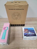 Boxed portable air conditioner fan next to smaller boxed handheld fan and fan humidifier on wooden surface