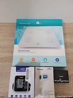 Lot showing all four devices boxed: 1 By One wireless smart scale, Cazon blood pressure monitor, Berrcom infrared thermometer, TUGER pulse oximeter.