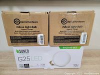 Two cardboard boxes labeled Basics Hardware Edison Vintage Light Bulbs ST64, one white box labeled Sunco Lighting G25 LED 10 pack.