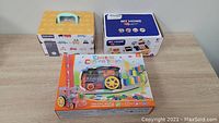 Three boxed toys: electric domino train with plastic dominoes, My Home cash register toy by Zovi model 3220A, and a box for construction vehicles toy.