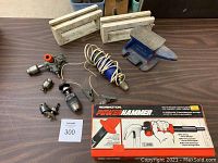Photo showing small blue metal bench anvil, boxed Remington Power Hammer with manual and powder loads, two drill chucks, a handheld drill attachment with red knob, a coiled corded electric tool, and miscellaneous metal parts on a table.