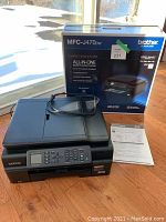 Brother MFC-J470dw printer displayed with its original box and owner's manual on wood floor near window.