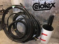 Photo showing multiple black plastic tubing coils of varying sizes leaning against a wall with reflective insulation, also a white container holding two plastic tubes.