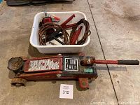 Photo showing the red hydraulic car jack and a white tub containing three sets of jumper cables with red clamps.