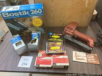 Photo of Bostik 260 electric glue gun, original box, Swingline stapler, glue sticks, and multiple packages of staples arranged on a table.
