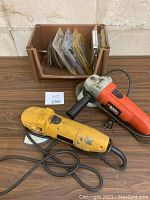 Overview photo showing DeWalt and Black & Decker grinders with sanding discs in brown storage container behind them on wooden table surface.