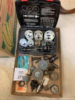 Open plastic case with multiple sizes of white hole saw cutting tips arranged inside along with arbors and pilot bits. Cardboard tray underneath contains additional drill accessories, mandrels, and saw blades.