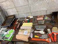 Overview of assorted painting tools including multiple paint rollers, paint brushes, scrapers, and a paint tray with dried paint.