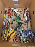 Multiple assorted pliers, scissors, and wire cutters in cardboard box showing condition and variety