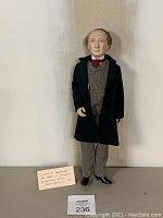 Full view of the Sir John A MacDonald porcelain doll standing upright wearing a dark coat, gray suit, and red tie.