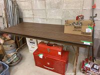Full view of the folding table with woodgrain top and metal legs in a storage area surrounded by boxes and various items.