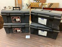 Four black plastic toolboxes stacked in two pairs on a wooden table. Each toolbox has a handle on top and two clasps for closure. The boxes show signs of wear and scratches, with labels indicating the size as 19 inches.