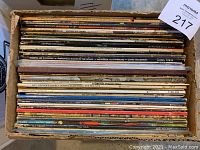 Cardboard box filled with vertical stack of vinyl LP sleeves showing varied spines