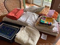 Photo showing pile of assorted vintage linens including embroidered white linen, white crocheted cloth, coral colored napkins, multicolored striped napkins, and printed floral textiles, plus a blue framed floral tray