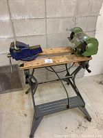 Wide view of workbench showing mounted vice on left and green bench grinder on right mounted to a wood plank top with metal framing base.