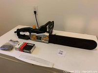 Full view of Yard Works electric chainsaw with chain guard on, additional chains in plastic bag, Oregon box on the table.